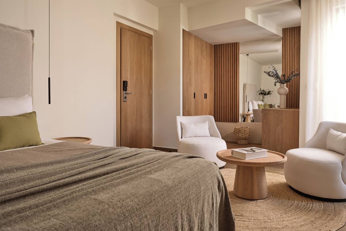 Une chambre d’hôtel moderne avec un grand lit, deux fauteuils blancs, une table basse en bois et une armoire en bois.