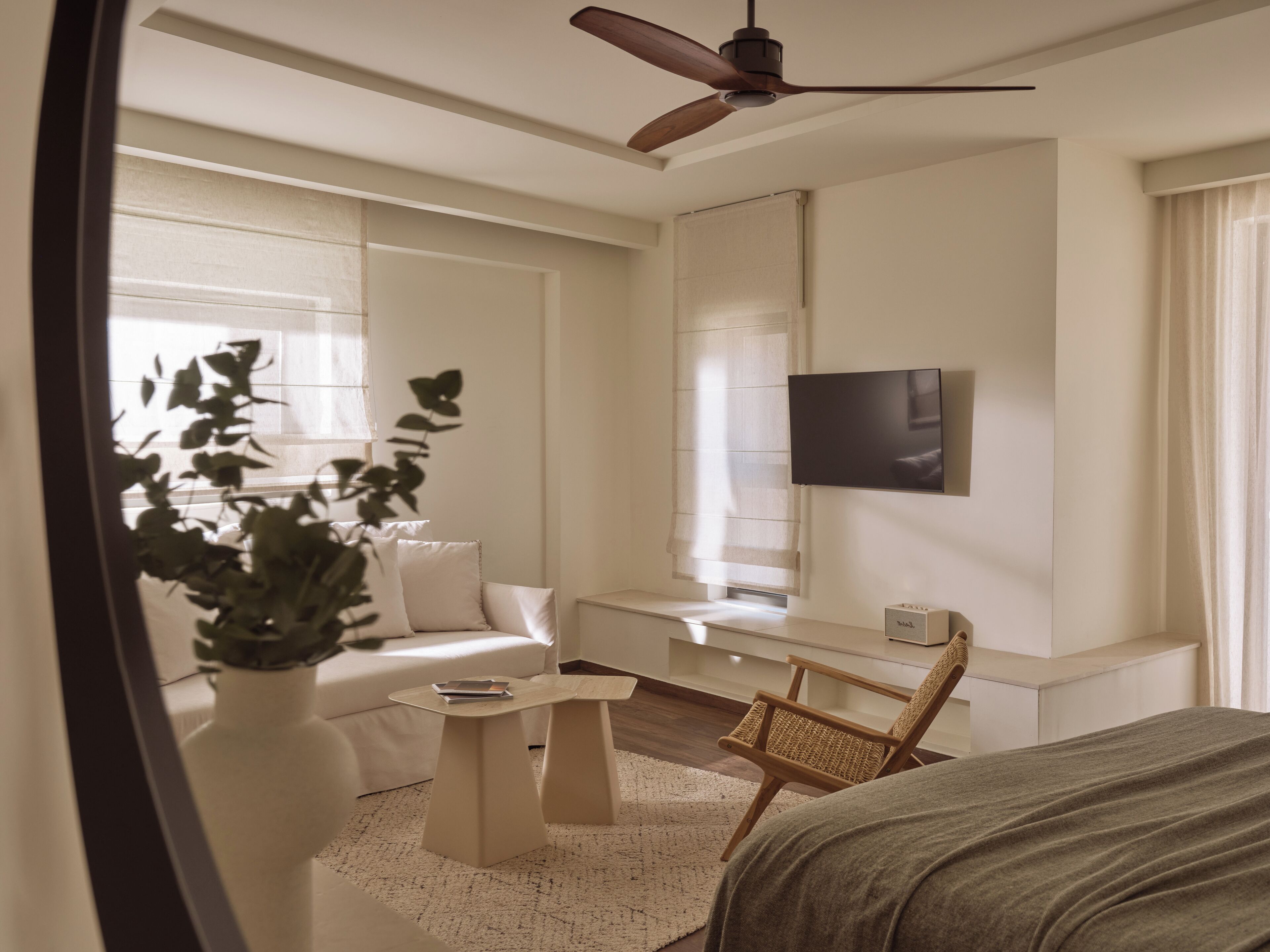 A modern hotel room with a bed, a flat-screen TV mounted on the wall, a ceiling fan, a seating area with a sofa and a chair, and a large window with curtains.