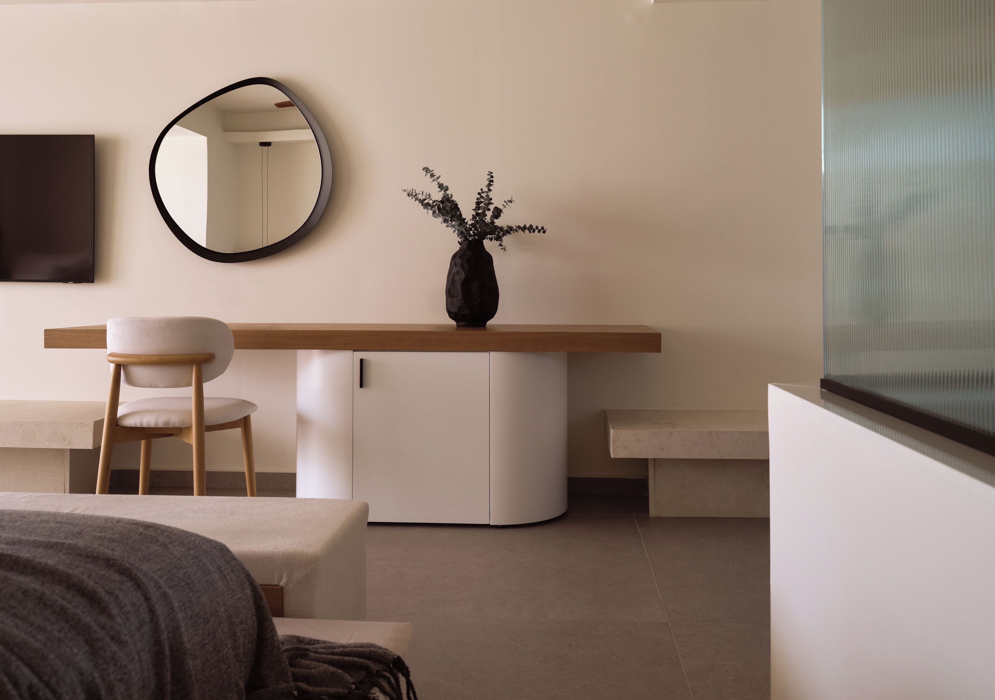A modern bedroom with a bed, a desk with a chair, a small cabinet, a round mirror, and a vase with flowers.