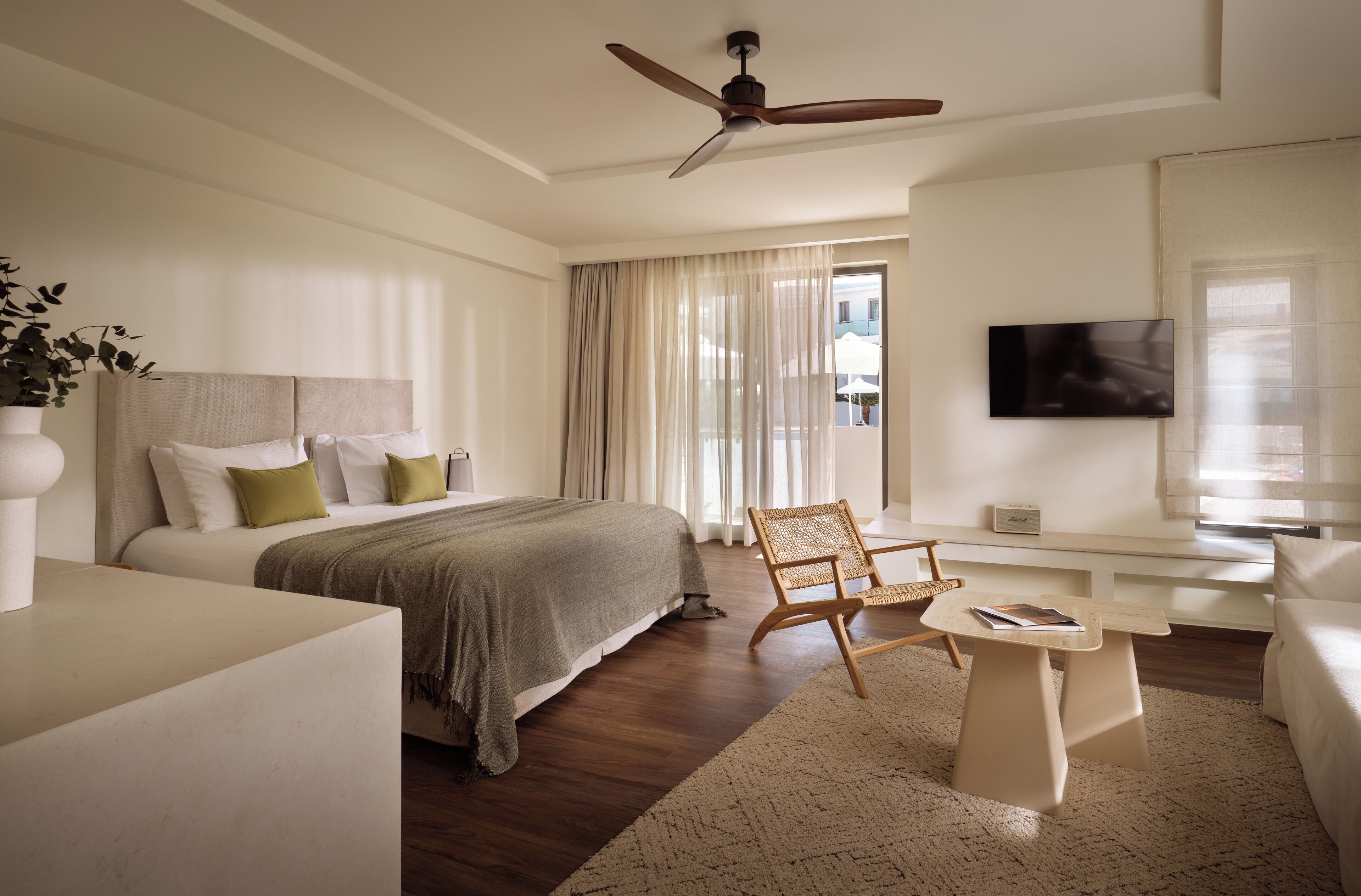 A modern hotel room with a bed, a chair, a small table, a ceiling fan, and a TV.