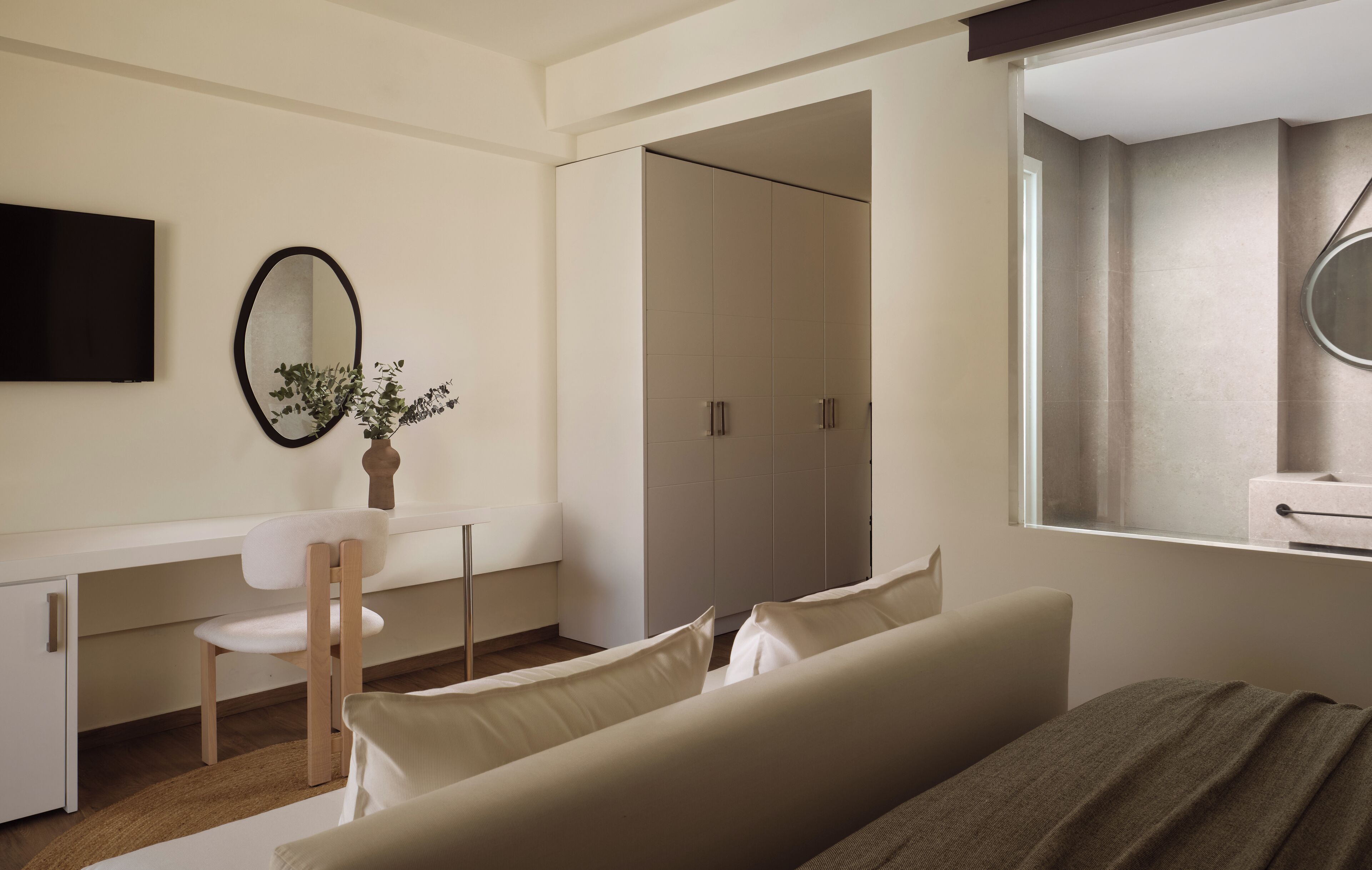 A modern hotel room with a bed, a desk with a chair, a large mirror, and a wardrobe.