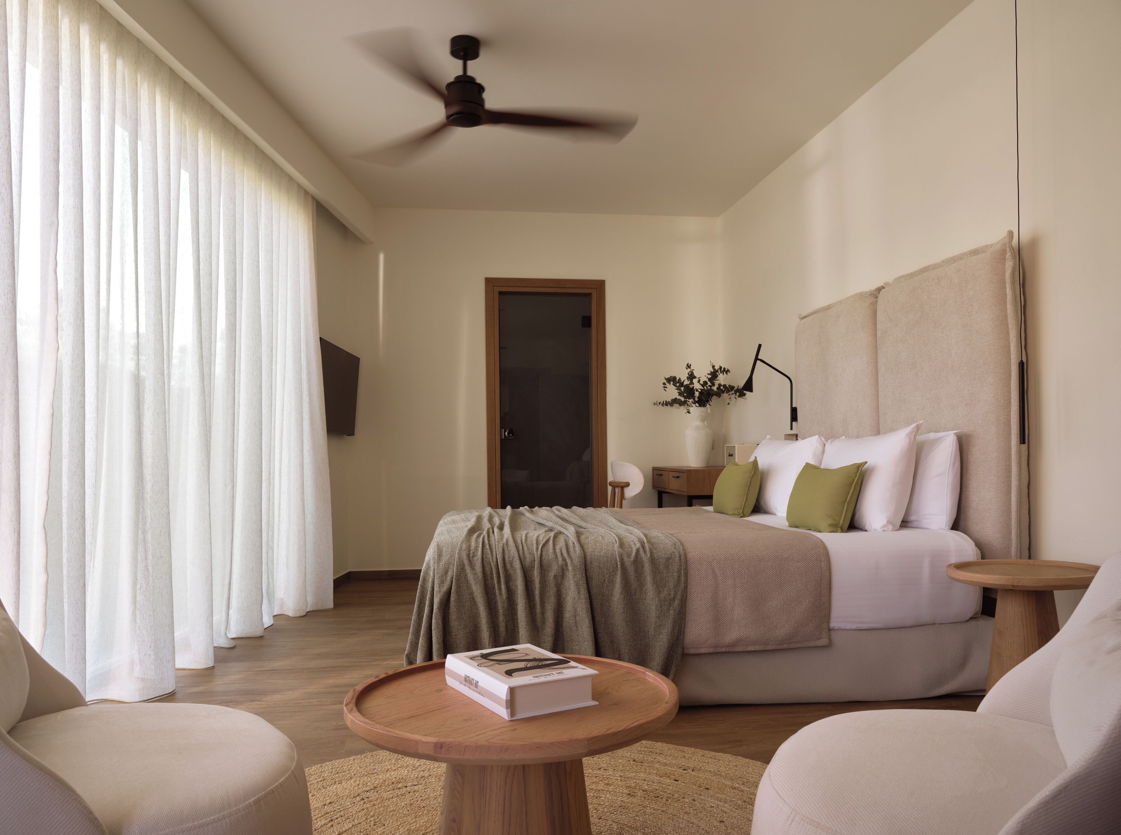 A modern hotel room with a bed, a round wooden table with a book, a ceiling fan, and a large window with curtains.