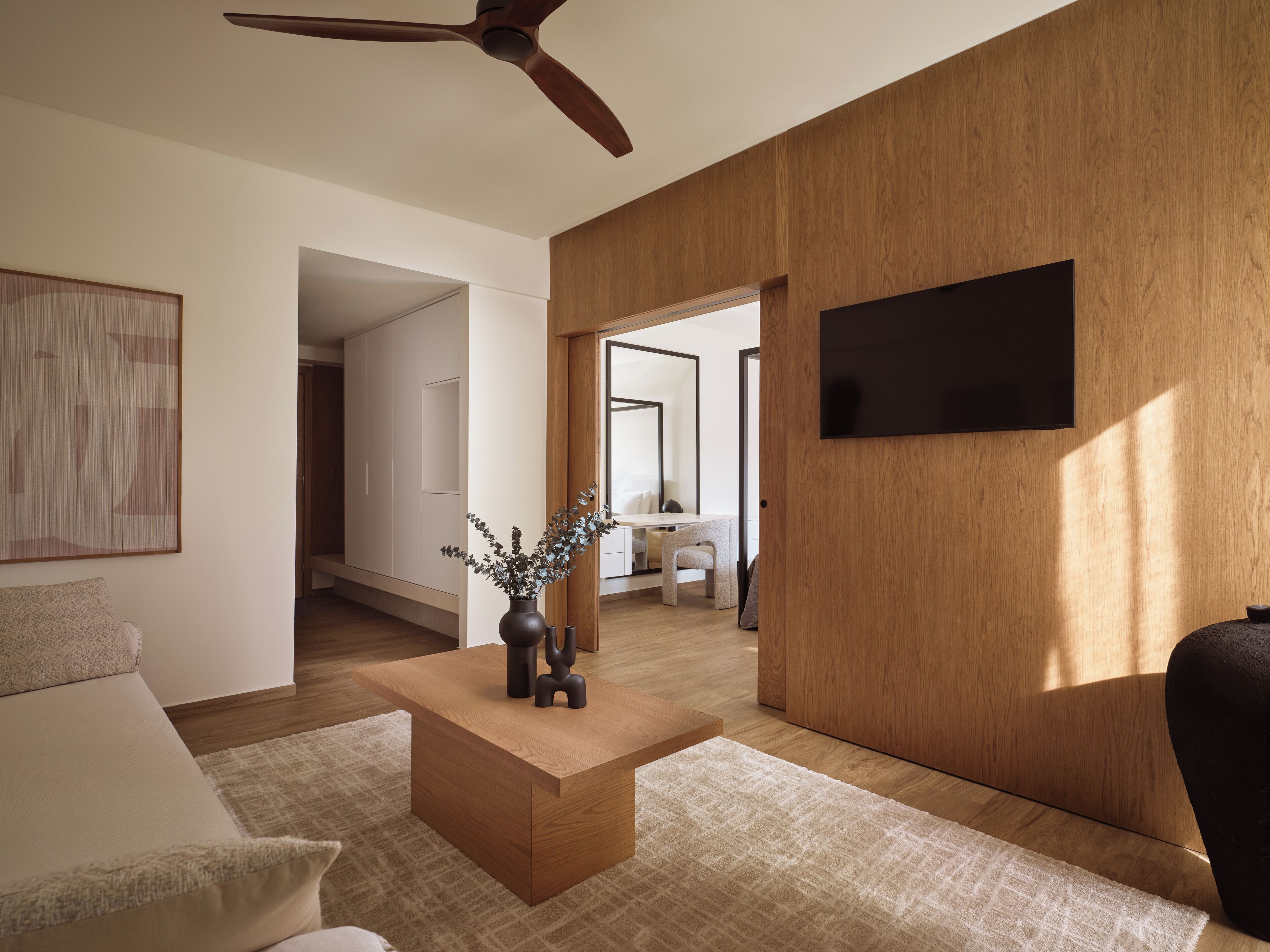 A modern living room with a wooden accent wall, a flat-screen TV mounted above a wooden cabinet, a coffee table with decorative items, and a ceiling fan.