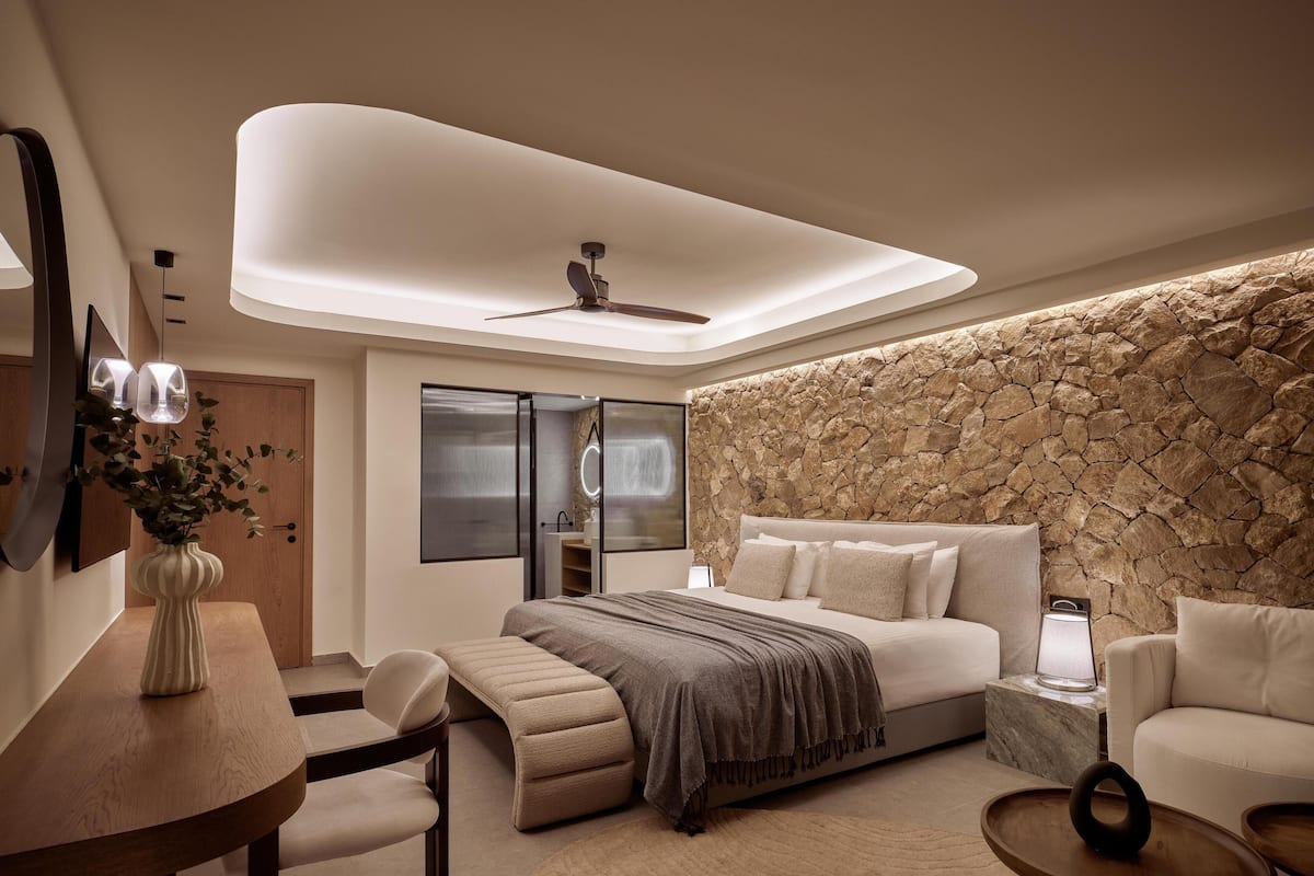 Une chambre moderne avec un mur d’accent en pierre, un lit recouvert d’une couverture grise, une table de chevet avec une lampe et un ventilateur de plafond.