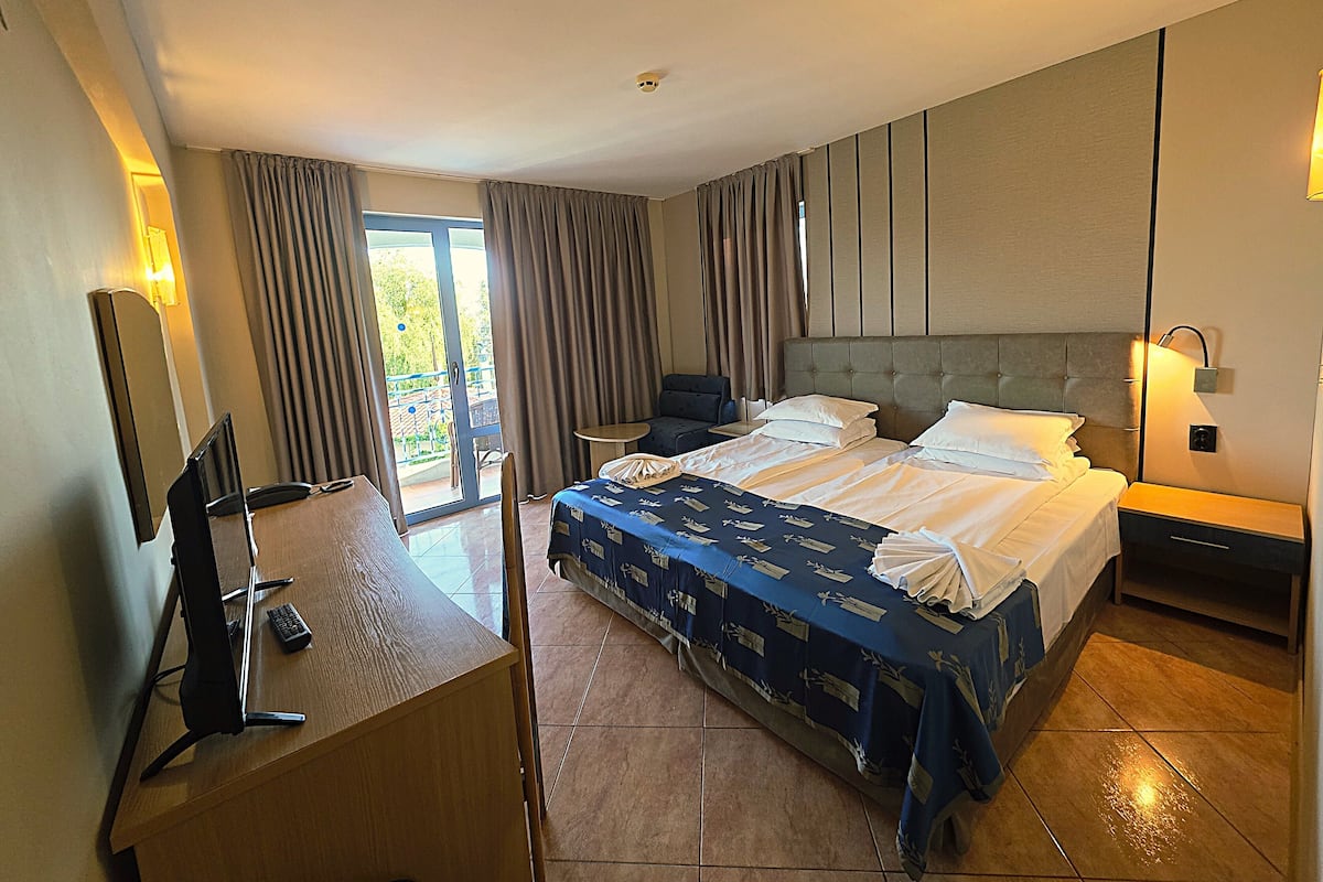 Hotellihuone, jossa on suuri sänky, työpöytä tuolilla, pieni pöytä ja parveke, jossa on verhot.