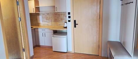 A compact kitchen with a white refrigerator, a microwave, and a sink. There is a wooden door and a bench in the corner.