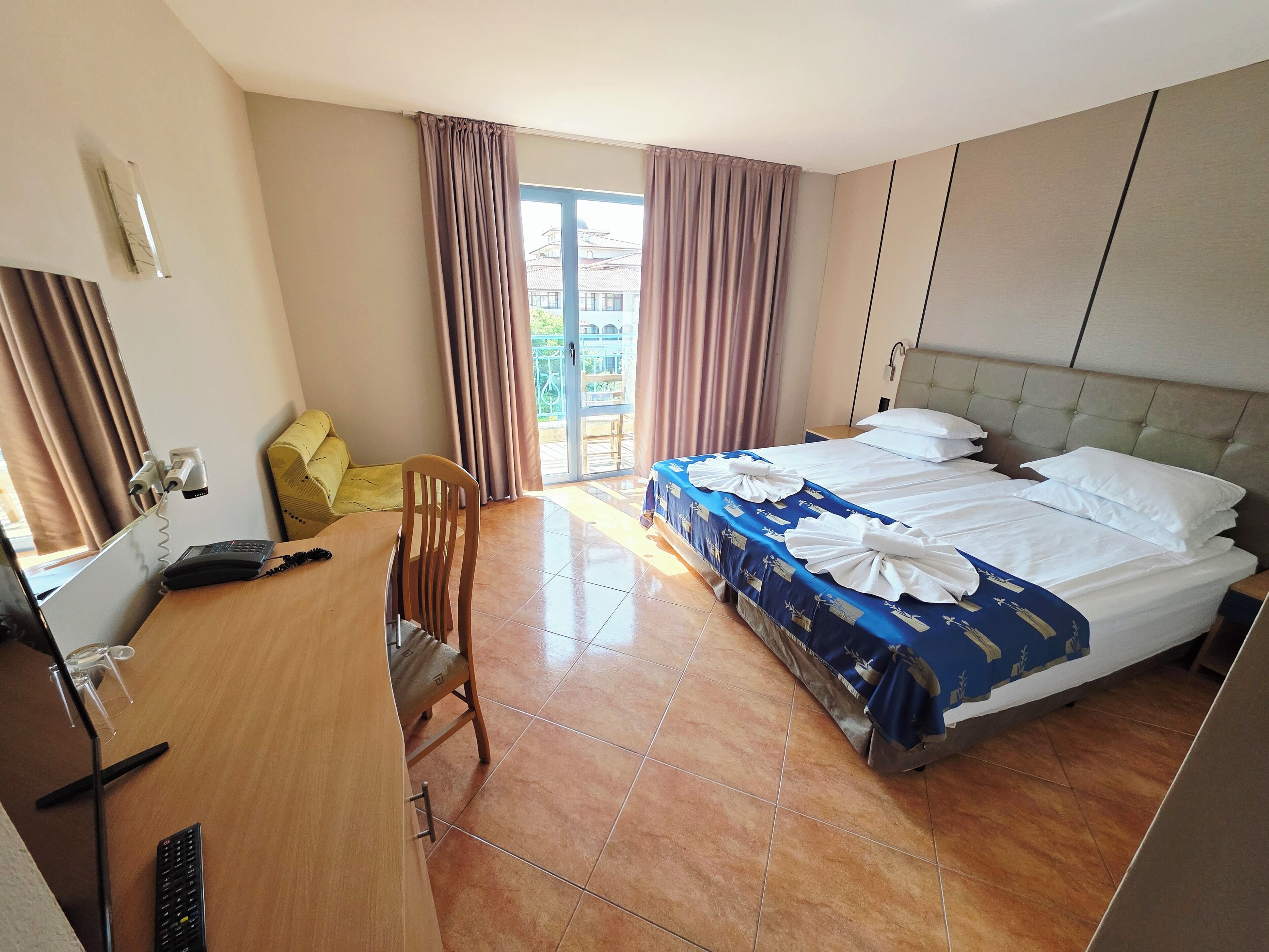 A hotel room with a bed, a desk with a telephone, a chair, and a window with curtains.