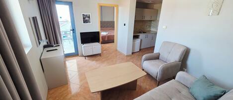 A living room with a sofa, a coffee table, and a TV. There is a kitchen area with cabinets and a sink.