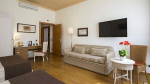 A furnished living room with a sofa, a chair, a small table with a plant, a television mounted on the wall, and a desk with a lamp. - Piazza Paradiso Accomodation (Siena)