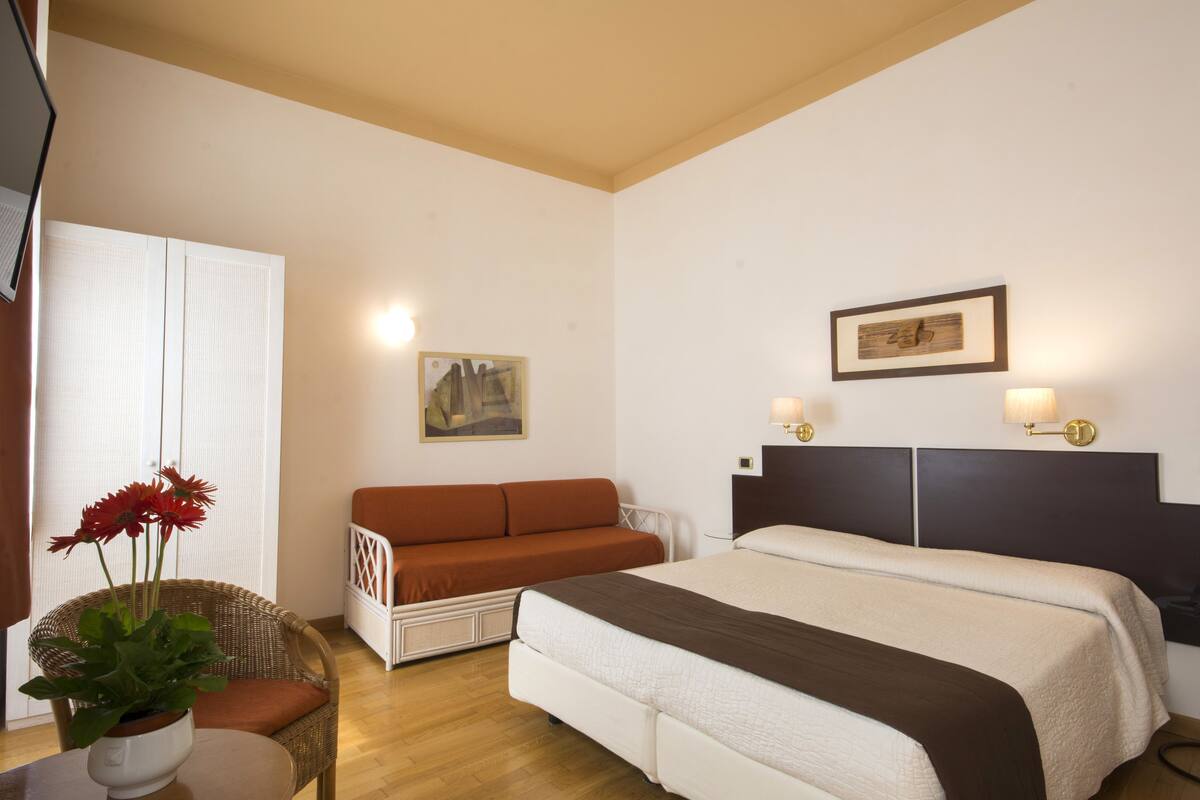 Une chambre d’hôtel avec un lit, un canapé, une chaise et une table sur laquelle se trouve un vase de fleurs.