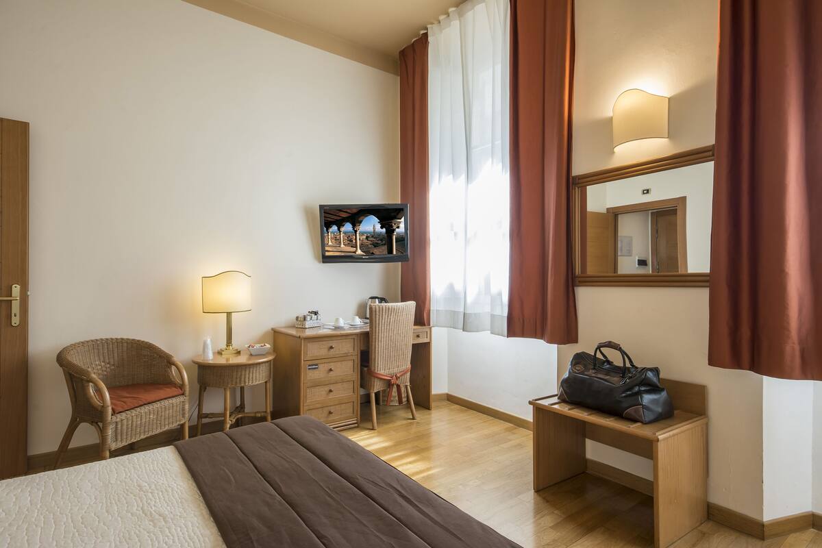 Une chambre d’hôtel avec un lit, un bureau, une chaise, une petite table avec une lampe, un miroir et une vue sur la ville par la fenêtre.