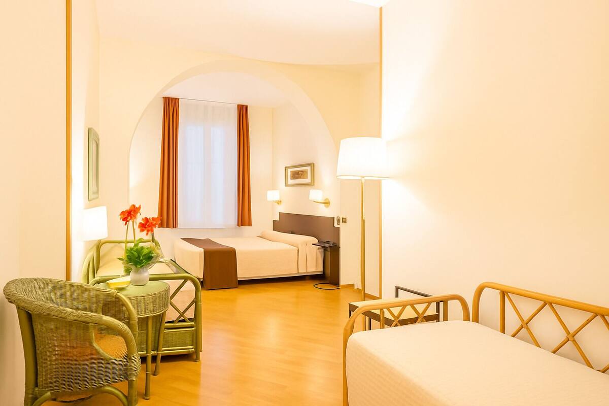 Une chambre d’hôtel avec un lit, un bureau avec une chaise, une lampe et une petite table agrémentée de fleurs.