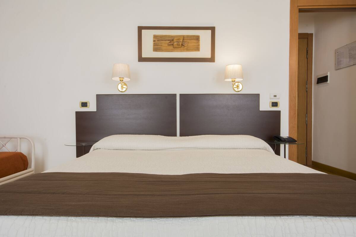 Une chambre d’hôtel avec un grand lit, deux tables de chevet, une petite table, un tableau encadré au mur et deux lampes fixées au mur.