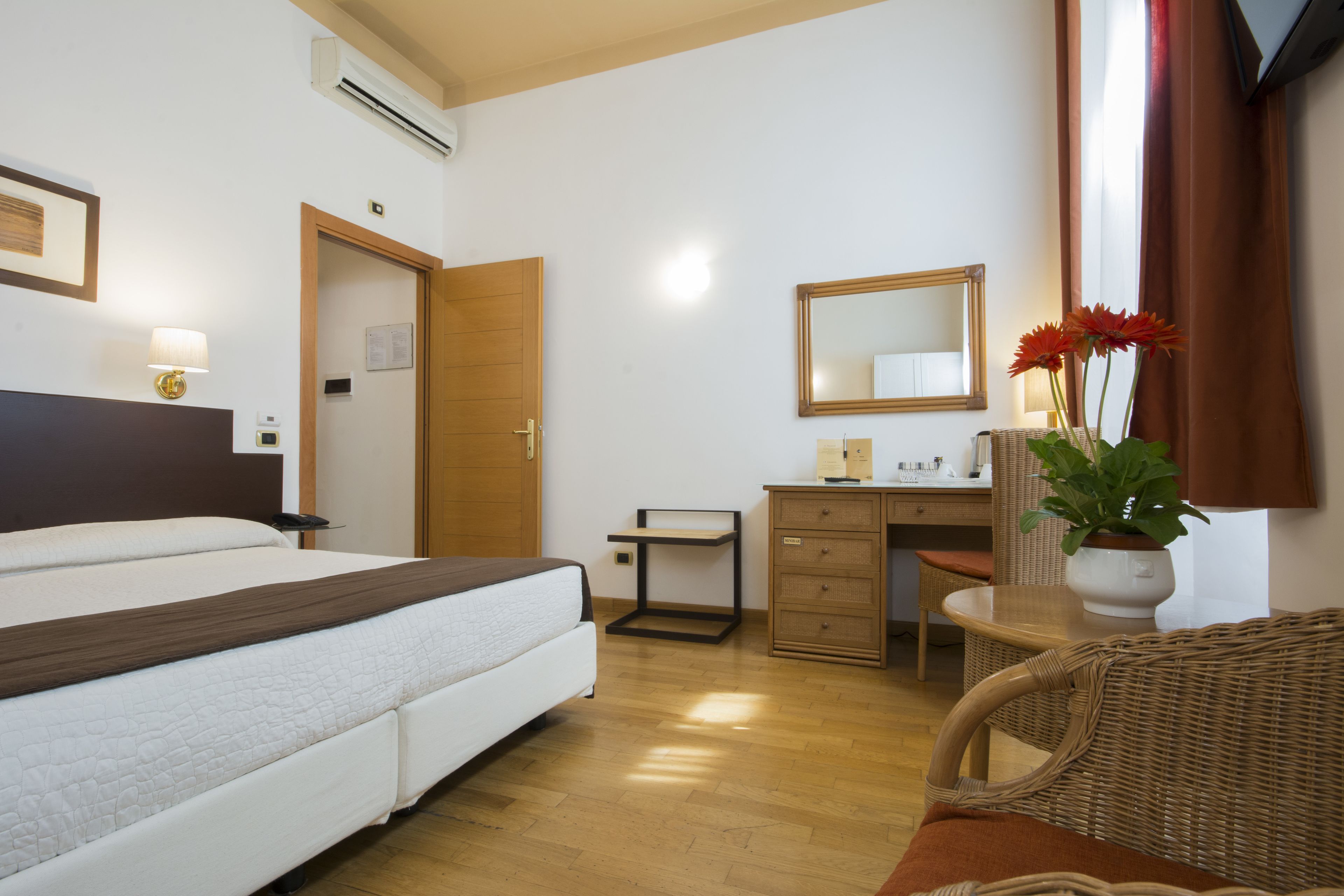 A hotel room with a bed, a desk, a chair, a dresser, a mirror, and a potted plant.