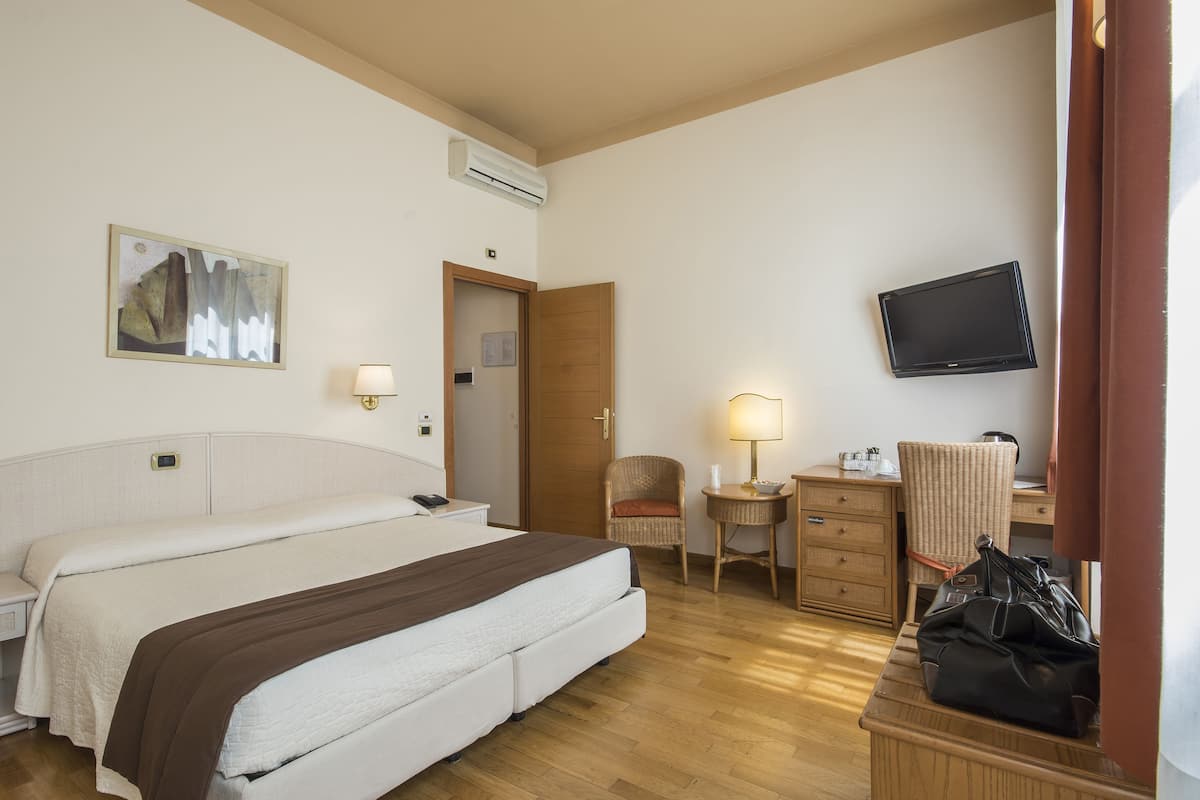 Une chambre d’hôtel avec un lit, une table de chevet, une lampe, une chaise, un bureau, une télévision et un sac posé sur le sol.