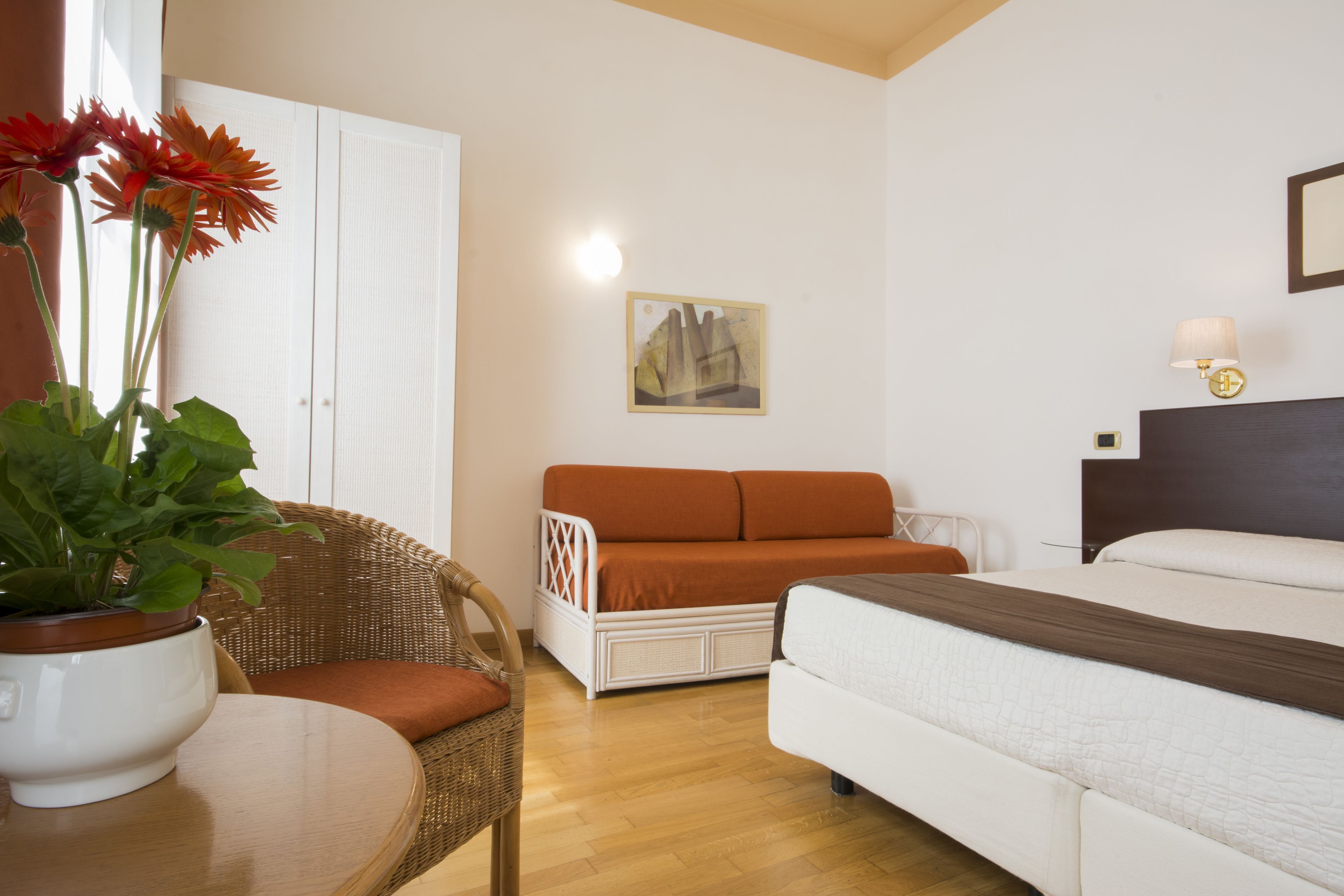 A hotel room with a bed, a sofa, a chair, a table with a potted plant, and a wall-mounted lamp.