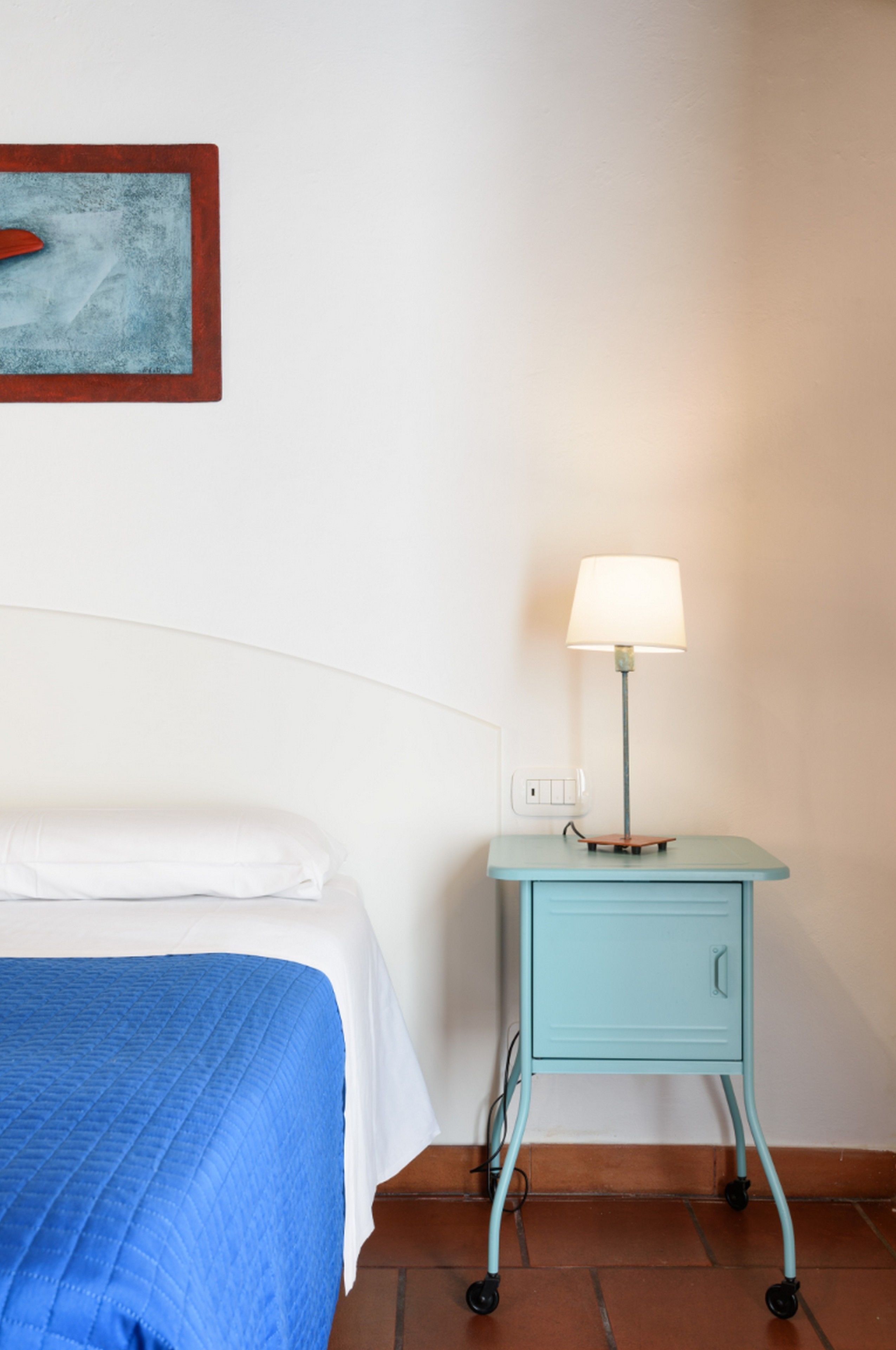 Un lit bien fait, recouvert d’une couverture bleue, une petite table de chevet turquoise avec une lampe, et une prise de courant fixée au mur.