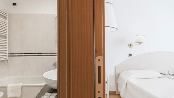 A modern hotel room with a wooden door, a white sink, a bathtub, a bed with white bedding, and a bedside table with a lamp. - Piazza Paradiso Accomodation (Siena)