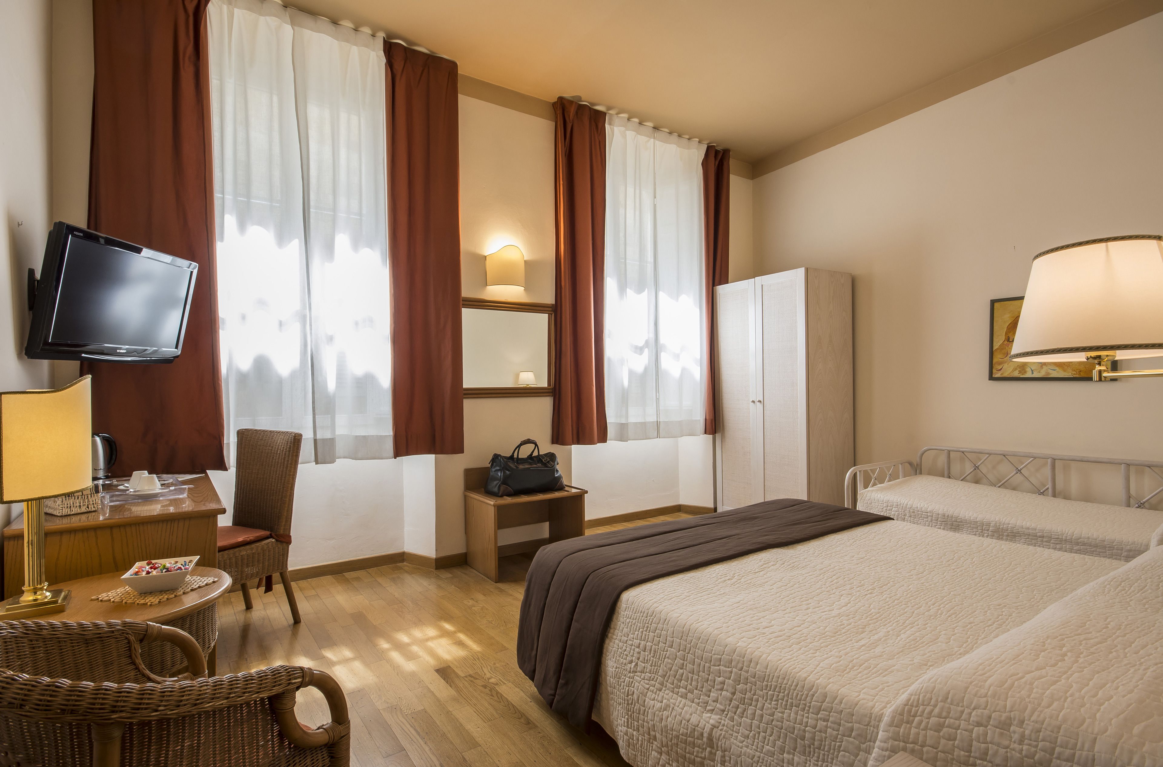 Una habitación de hotel con cama, escritorio con televisor, una silla, lámpara y armario.