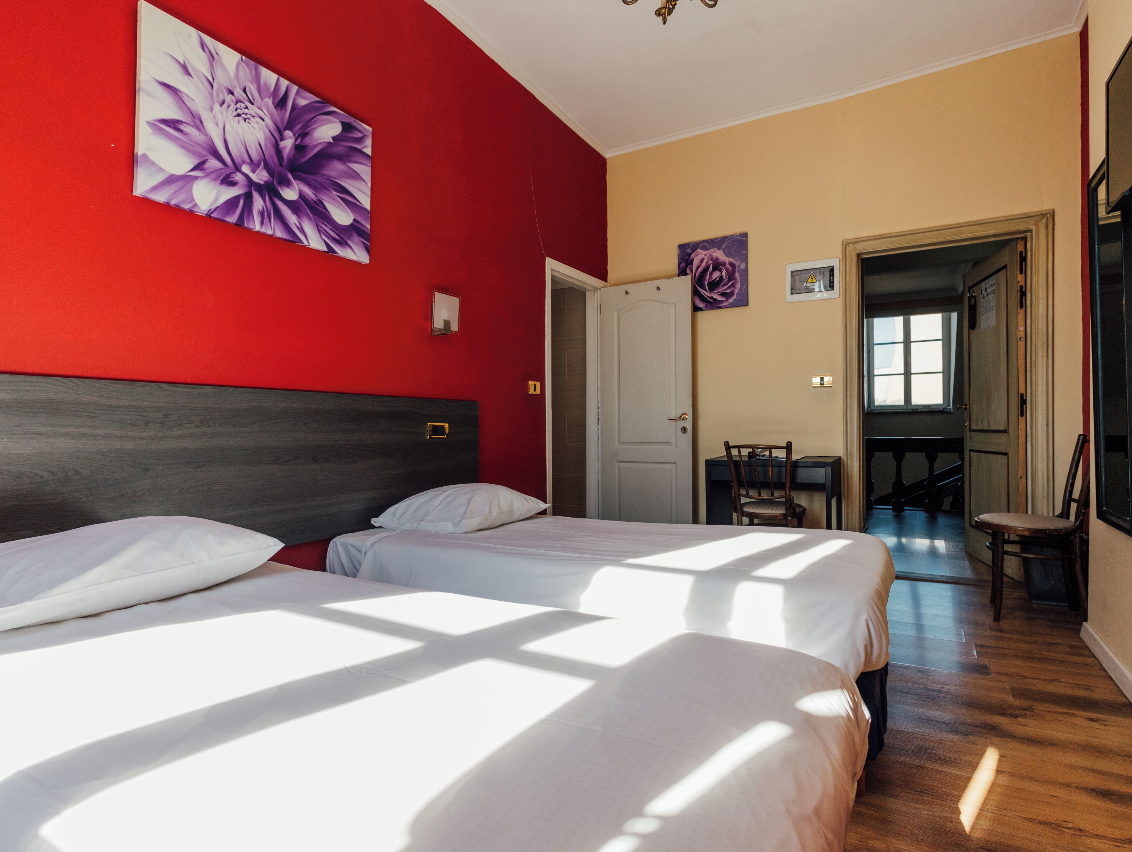 Habitación de hotel con dos camas, cabecera de madera, pared roja y puerta que da a un comedor.