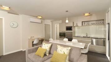 A modern living room with a grey sofa, a dining table, and a kitchen area with white cabinets and stainless steel appliances.