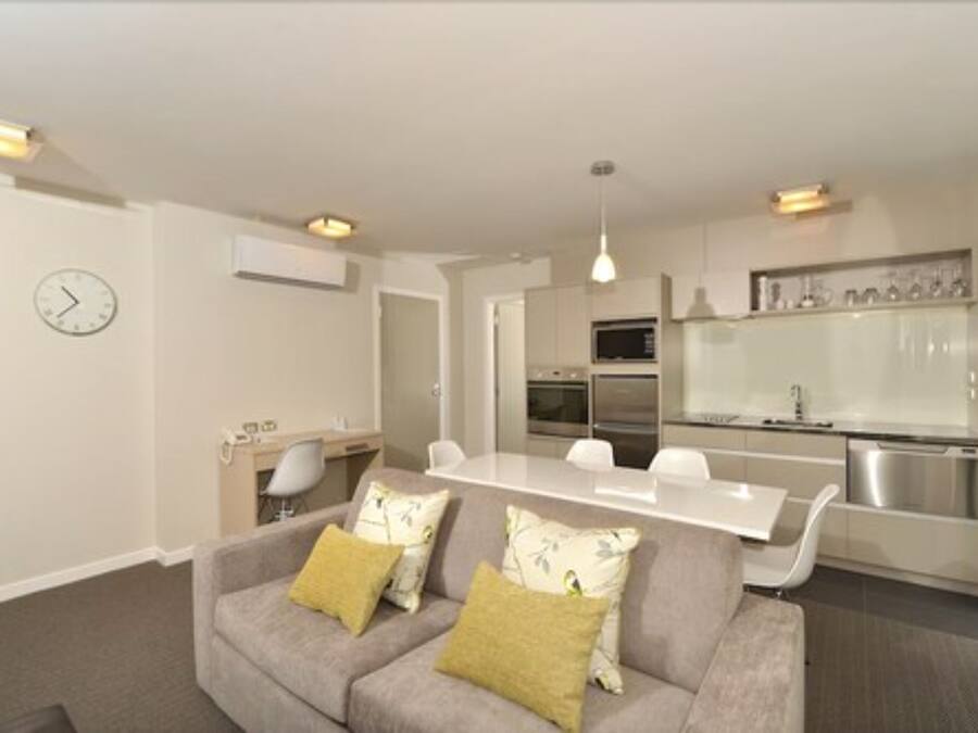 A modern living room with a grey sofa, a dining table, and a kitchen area with white cabinets and stainless steel appliances.
