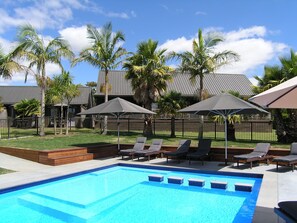 A swimming pool with lounge chairs and umbrellas, surrounded by palm trees and a fenced area. - Kerikeri Homestead Motel & Apartments (Kerikeri)