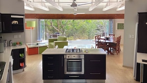 A modern kitchen with an island, built-in oven, and a dining area with a view of trees. - Bushwillow Collection (Hluhluwe)