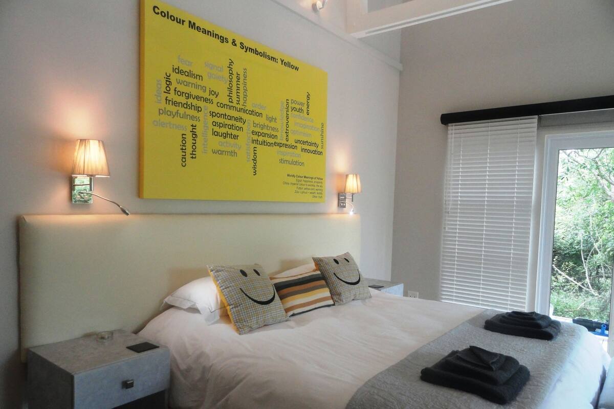 A neatly made bed with pillows, a bedside table, a wall-mounted lamp, and a window with blinds.