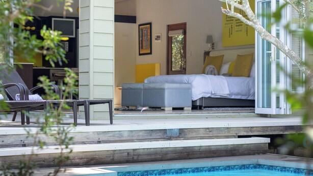 A poolside area with a bed and a chair, a small table, and a window with a view of the pool. - Bushwillow Collection (Hluhluwe)