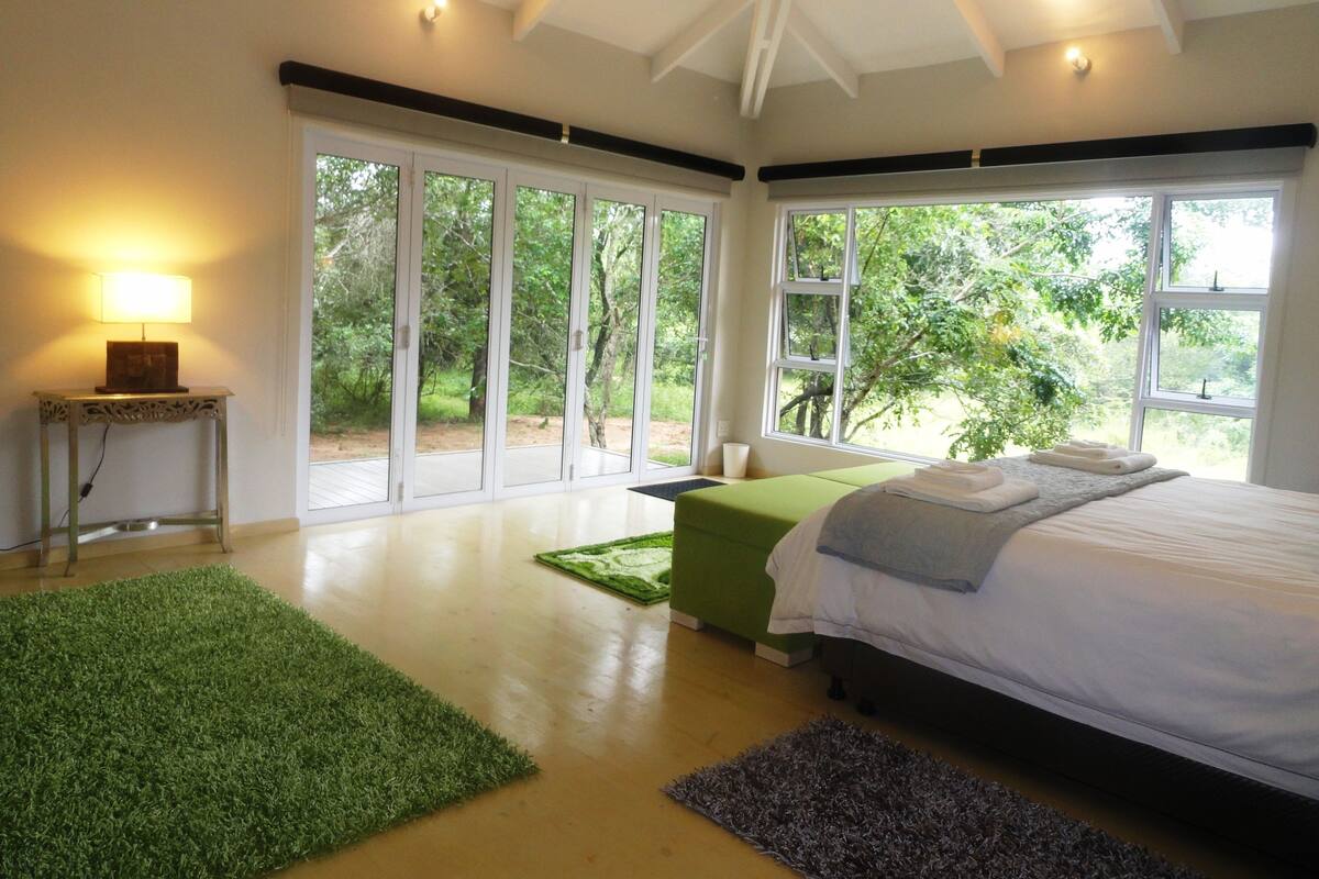 A bedroom with a large bed, a green bench, a small table with a lamp, and large windows with a view of greenery.