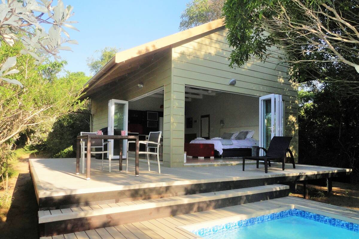 A poolside area with a wooden deck, a table with chairs, and a bedroom visible through an open door.