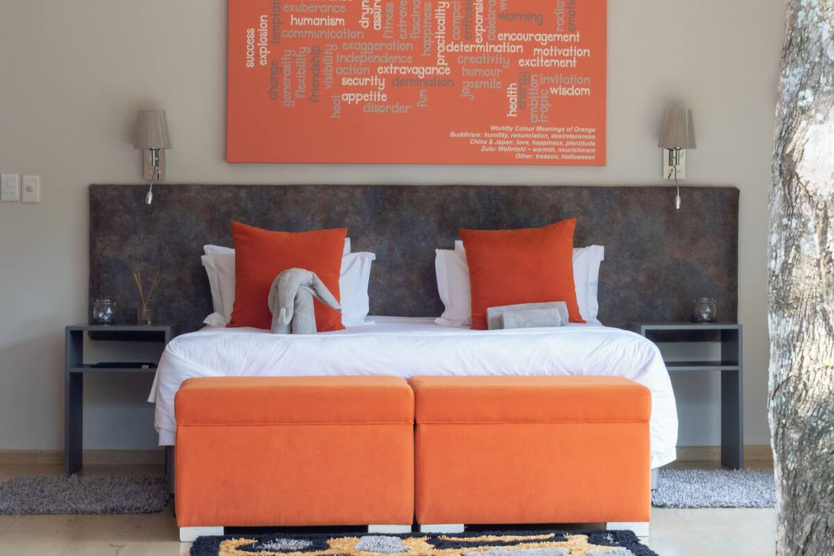 A bedroom with a large orange wall art piece listing color meanings and symbolism, a bed with white linens and orange pillows, and a nightstand with a lamp.
