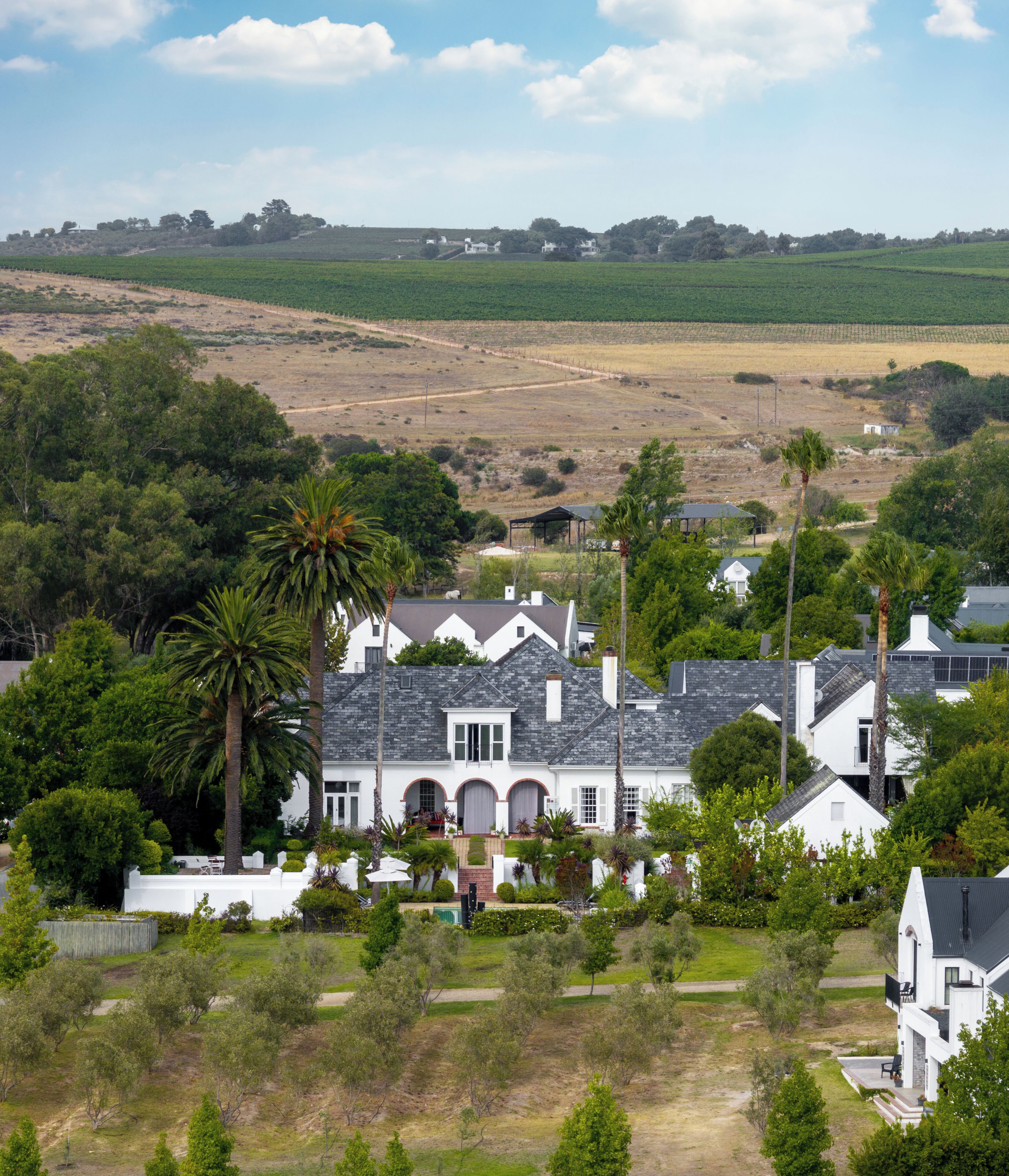 A large white house with a grey roof, surrounded by greenery and a hilly landscape.