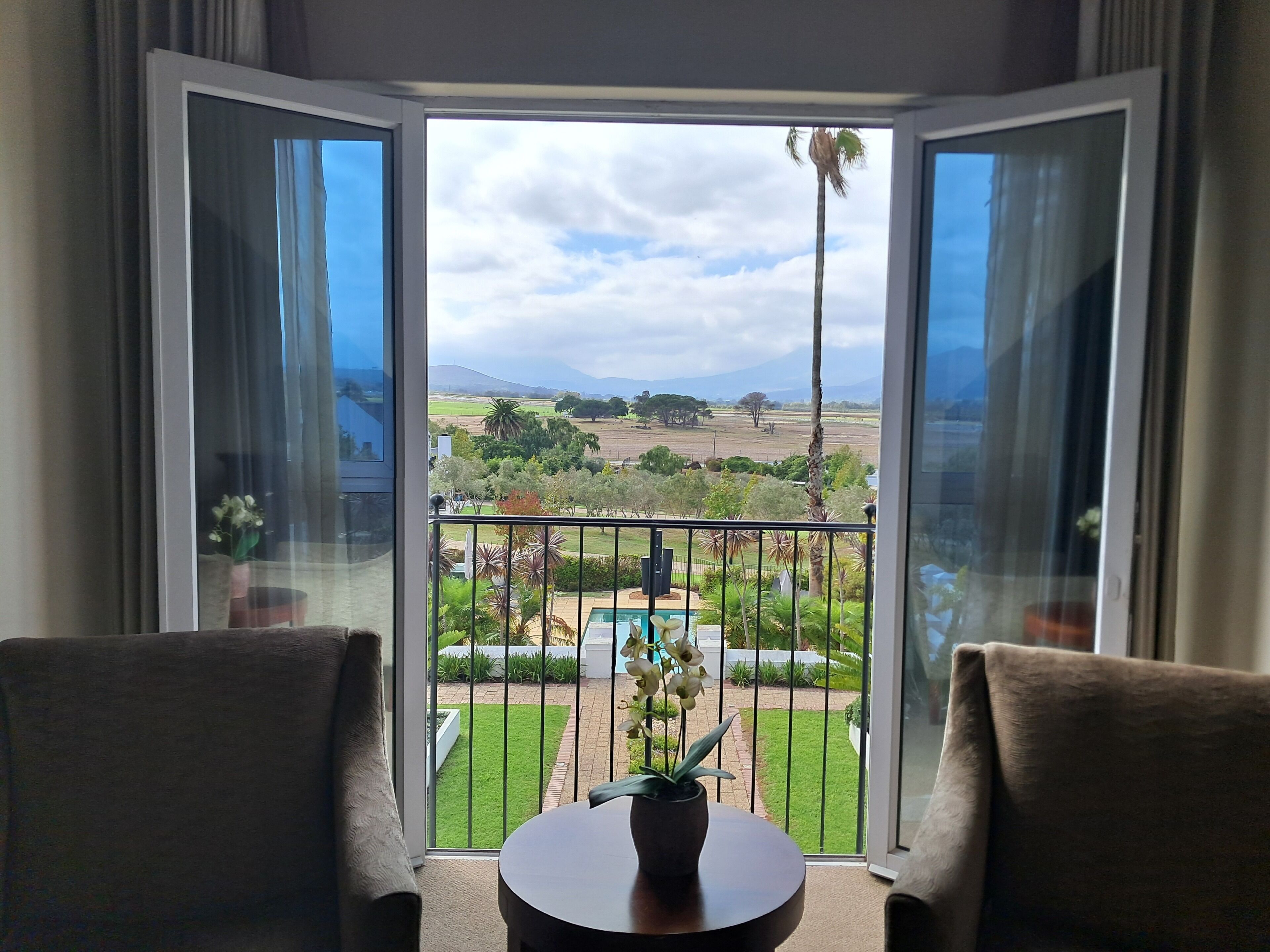 Ein Zimmer mit zwei Sesseln, die einen Balkon mit Blick auf einen angelegten Garten und ferne Hügel zeigen.