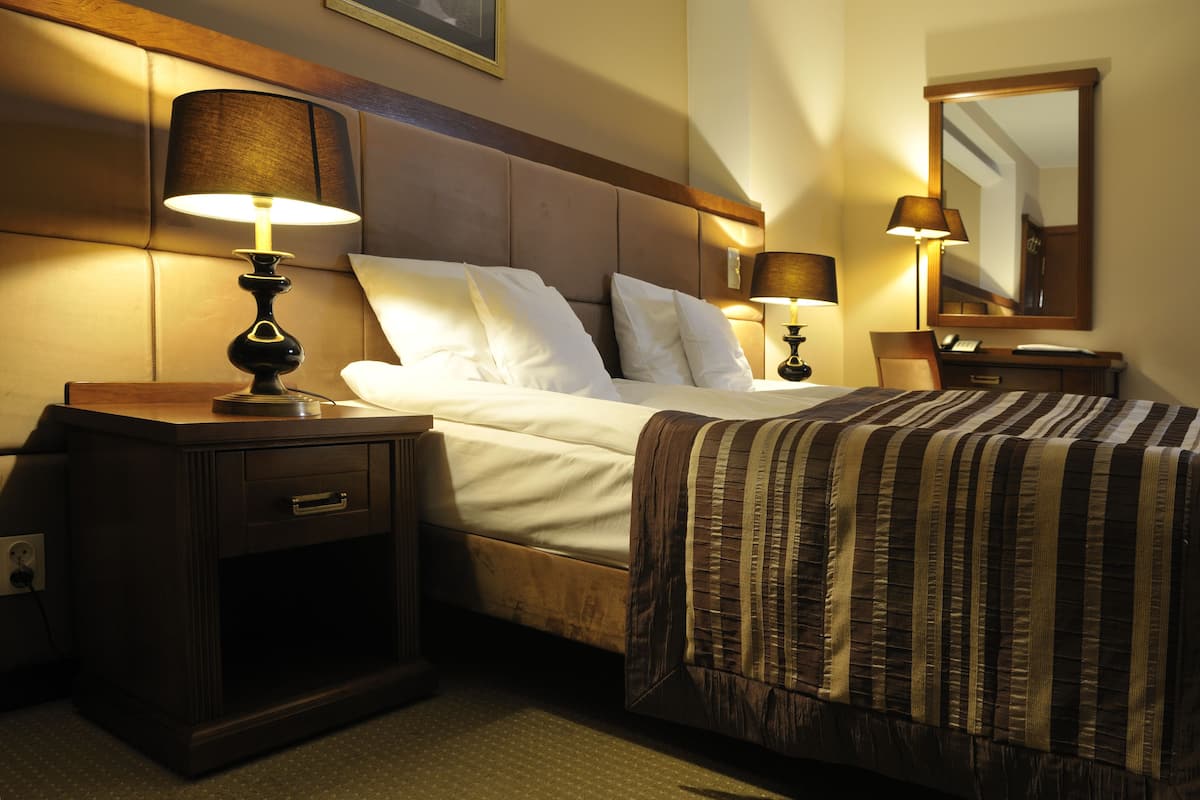 A hotel room with a bed, two bedside tables with lamps, a nightstand, a mirror, and a carpeted floor.