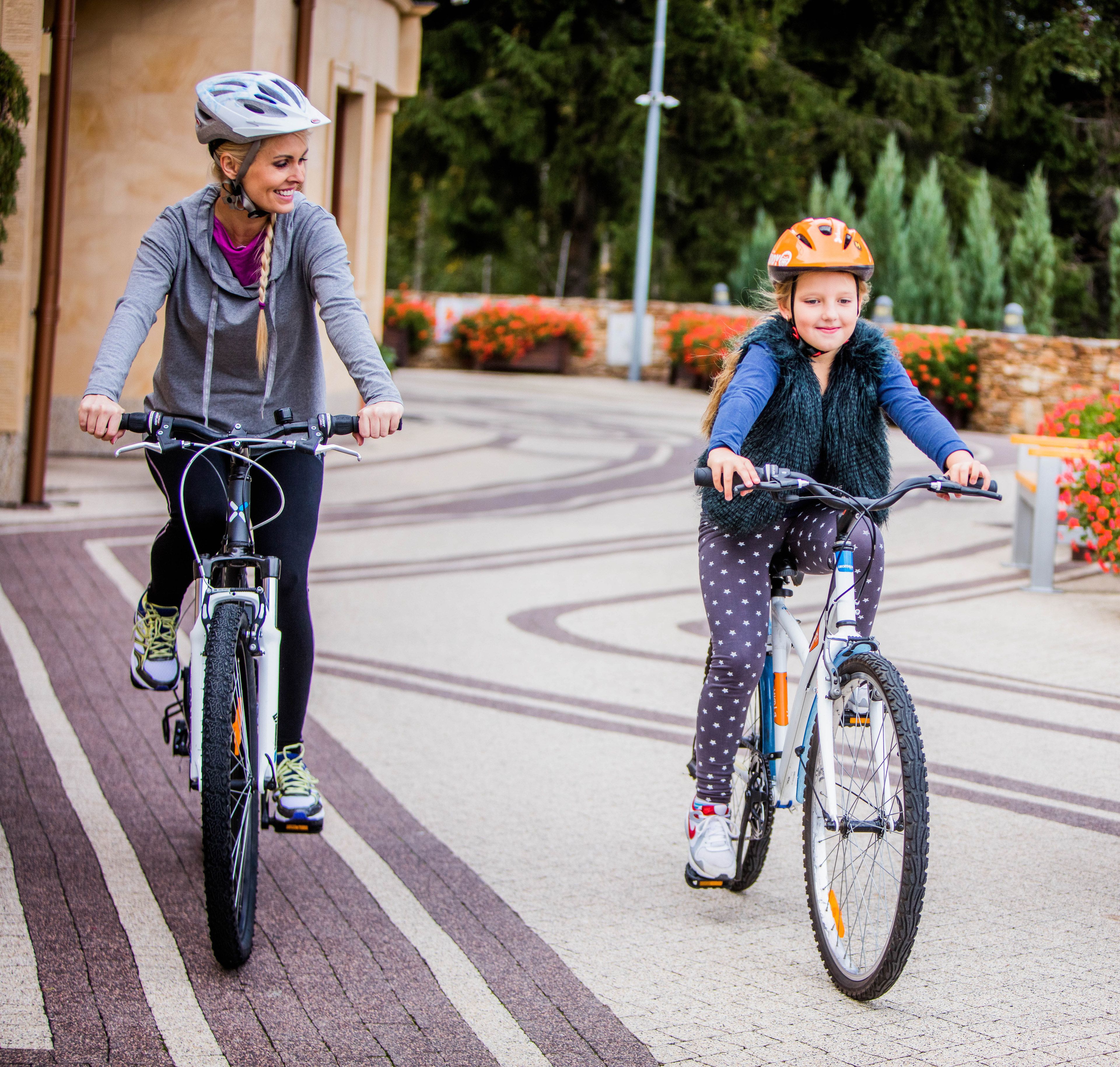 Two people riding bicycles on a paved path with a stone wall and greenery in the background.