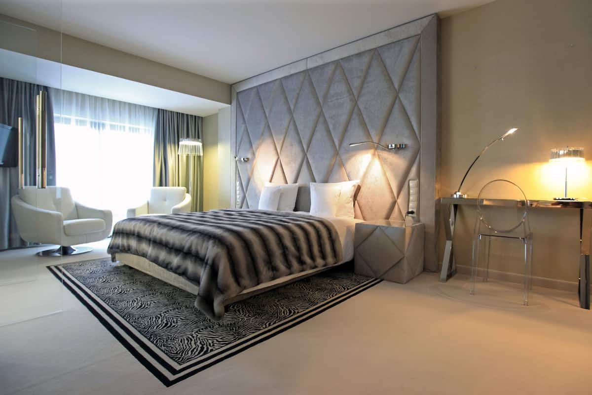 A modern hotel room with a large bed, a seating area with two chairs, a TV, and a decorative wall panel.