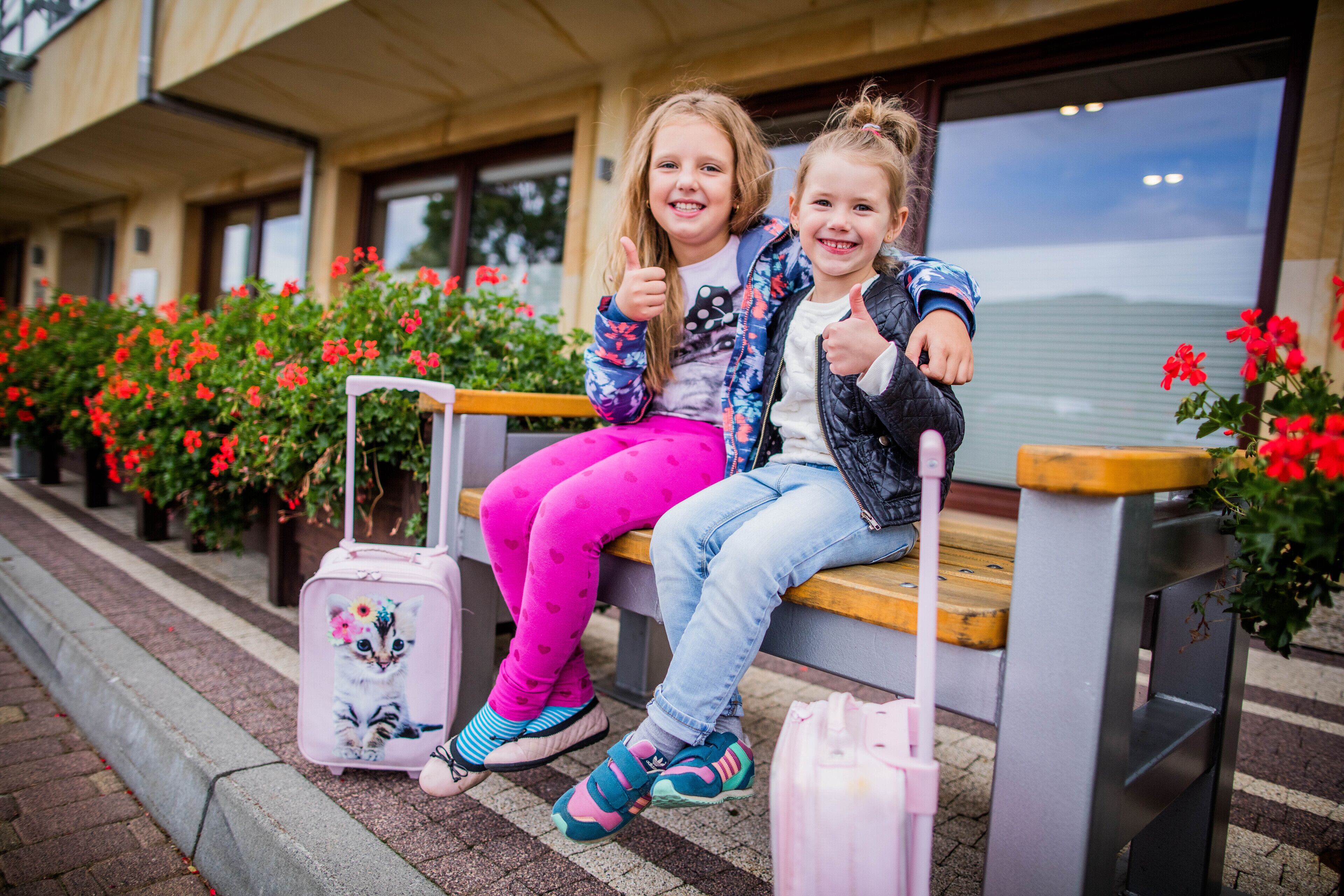 Two children sitting on a bench with a suitcase and a stroller nearby.