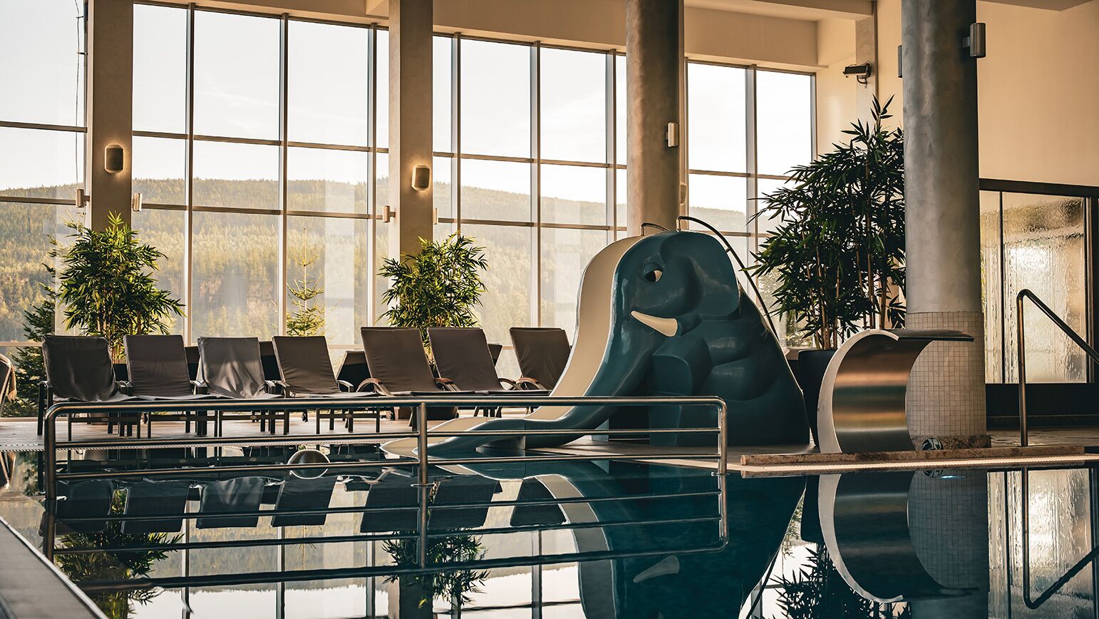 An indoor swimming pool with a water slide, lounge chairs, and large windows offering a view of a forested landscape.