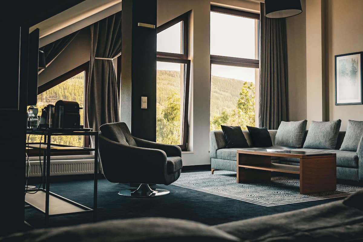 A modern living room with a sofa, armchair, and coffee table, large windows with curtains, and framed pictures on the wall.
