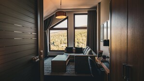 Ett modernt hotellrum med ett stort fönster som vetter mot en skog, en soffa med kuddar, ett litet bord och ett skrivbord med en lampa.