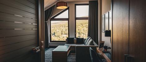 A modern hotel room with a large window offering a forest view, a sofa with cushions, a small table, and a desk with a lamp.