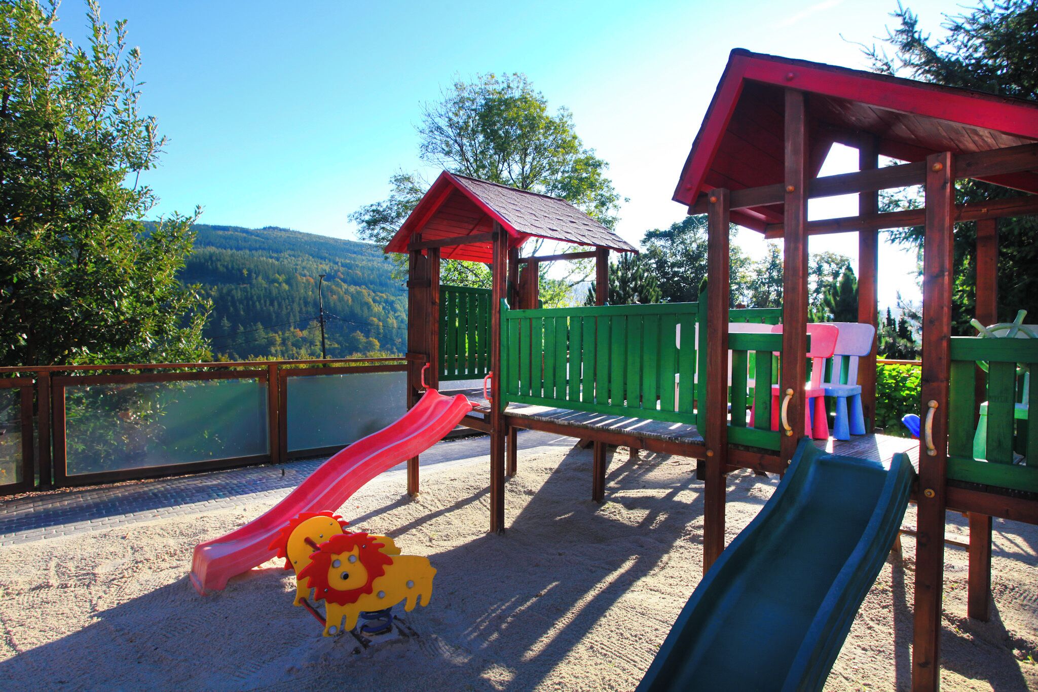 A playground with two slides, a wooden playhouse, and a view of a lake and forest.