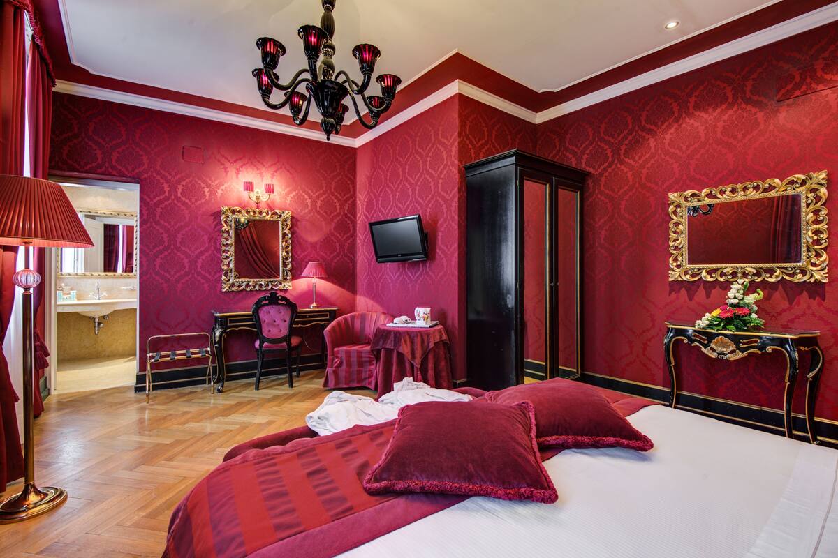 Ein Hotelzimmer in Rot gehalten, mit Bett, Schreibtisch, Stuhl, Fernseher und Spiegel.