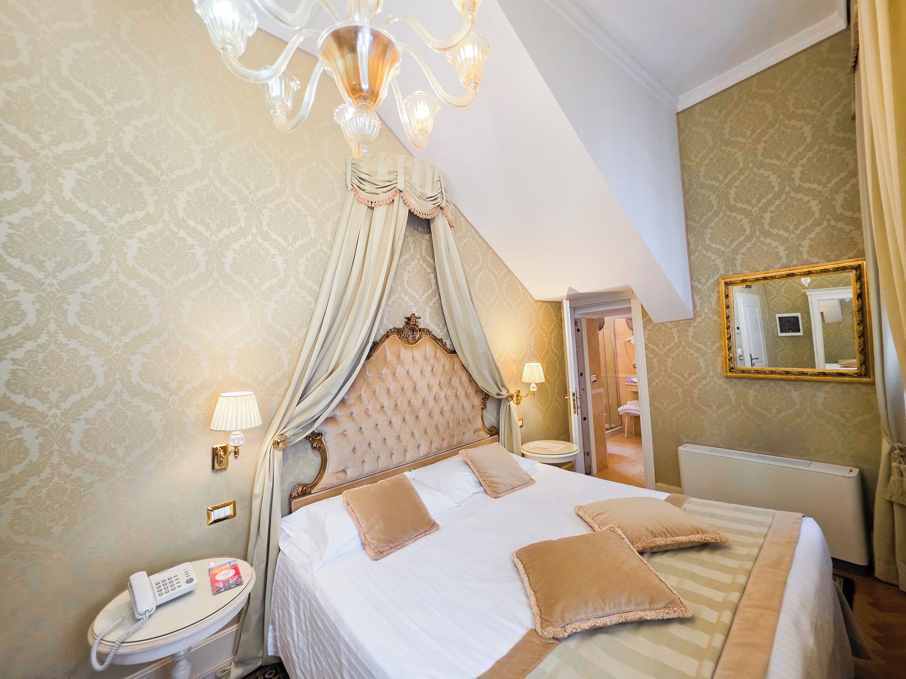 A bedroom with a four-poster bed, a chandelier, a bedside lamp, a telephone, and a mirror on the wall.