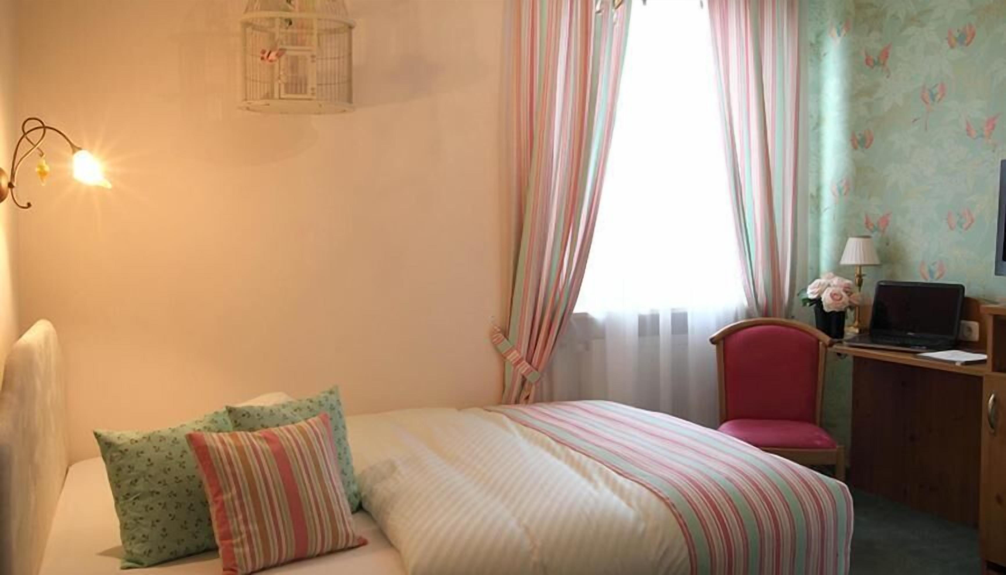Un dormitorio con cama, una silla, un escritorio con una computadora portátil, una lámpara y una ventana con cortinas.