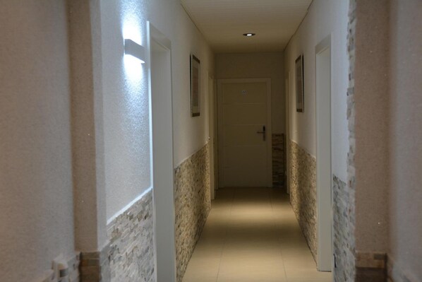 A narrow hallway with a tiled floor and walls with a stone-like texture. There are doors on both sides, and the hallway is well-lit with ceiling lights. - Alpina Hotel (Rosenheim)