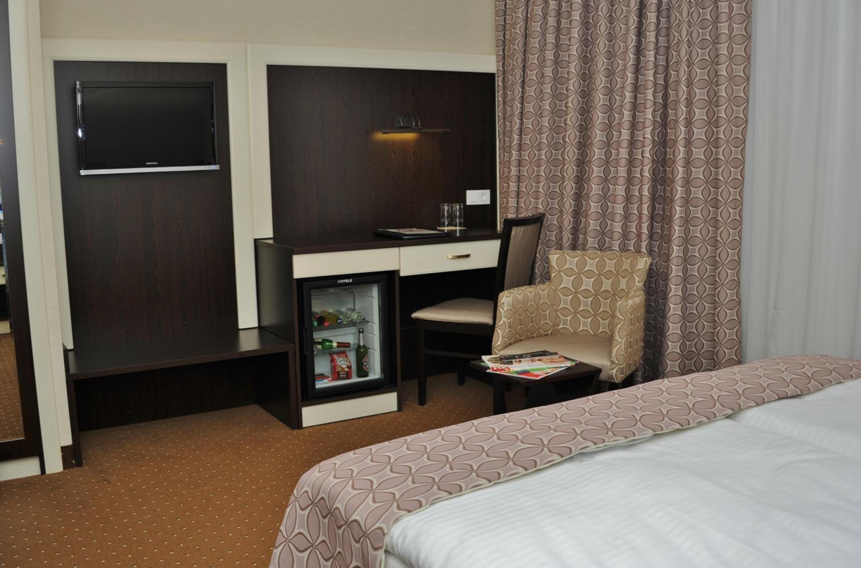 A hotel room with a bed, a desk with a minibar, a chair, and a television.