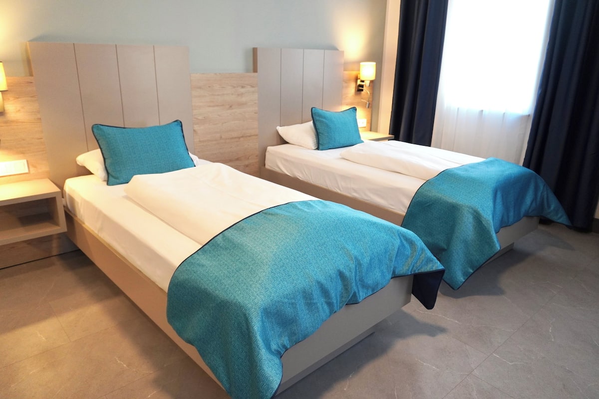 A hotel room with two beds, each with a blue bedspread and pillow, a wooden headboard, a nightstand with a lamp, and a window with curtains.
